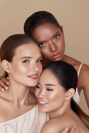 Diverse. Models Beauty Portrait. Ethnic Women With Nude Makeup And Glowing Skin Standing Together Against Beige Background. Caucasian, Asian And Mixed Race Female Hugging And Looking At Camera.の写真素材