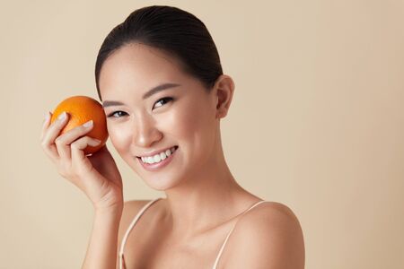 Woman And Fruit Beauty Portrait. Smiling Asian Model With Orange Looking At Camera Against Beige Background. Natural Vitamin C As Detox Treatment For Healthy, Smooth And Glowing Skin.の写真素材