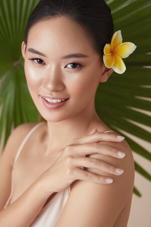 Beauty. Model Near Palm With Flower In Hair. Tender Asian Woman Touching Shoulder Portrait On Tropical Plant Background. Beautiful Girl Enjoying Hydrated Skin After Using Natural Organic Cosmetic.の写真素材
