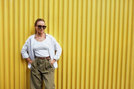 Woman. Portrait Of Sexy Model. Fashion Girl In Casual Clothes And Sunglasses Standing Against Yellow Metal Fence. Female In White Shirt And Top Tank Keeping Hands On Hips. Urban Lifestyle.の写真素材