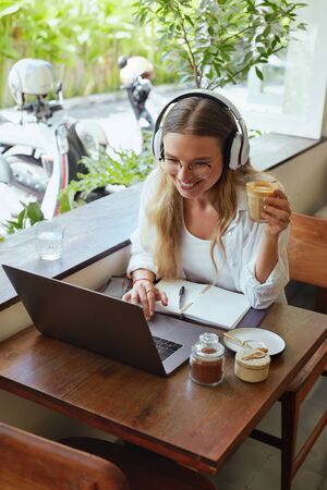 Girl At Coffee Shop. Woman Drinking Latte With Laptop And Headphones At Cafe. Beautiful Model In Glasses Working Or Studying Remotely At Comfortable Workplace On Vacation.の写真素材