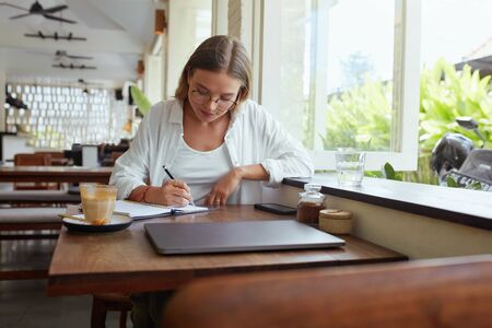 Business. Woman At Coffee Shop Writing In Planner Book. Beautiful Model In Glasses Enjoying Summer Vacation And Working. Online Education Or Modern Creativity Job At Comfortable Workplace.の写真素材