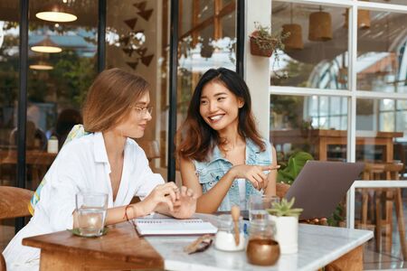 Girls At Coffee Shop. Women Having Call On Laptop At Cafe. Beautiful Colleagues In Casual Clothes Chatting At Comfortable Workplace On Summer Vacation. Modern Technologies For Digital Nomad Lifestyle.の写真素材