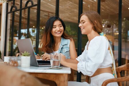 Women Working On Laptop At Cafe. Beautiful Ethnic Girls In Casual Clothes Having Discussion About Business On Terrace. Digital Nomad Lifestyle For Comfortable Work At Coffee Shop.の写真素材