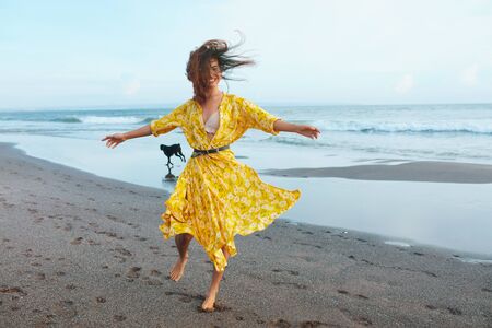Summer Fashion. Woman In Bohemian Clothing Walking With Dog On Beach. Beautiful Model In Maxi Dress With Floral Pattern Enjoying Resting On Ocean Shore. Boho Style For Fashionable Look On Resort. の写真素材