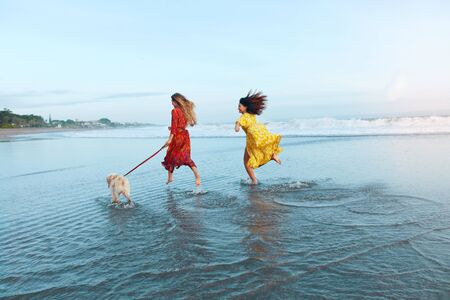 Girls With Dog On Beach. Running Women In Maxi Dresses Along Ocean Shore. Female Models In Trendy Outfit On Summer Vacation. Bohemian Clothing Style For Fashion Look On Dog-Friendly Resort.の写真素材