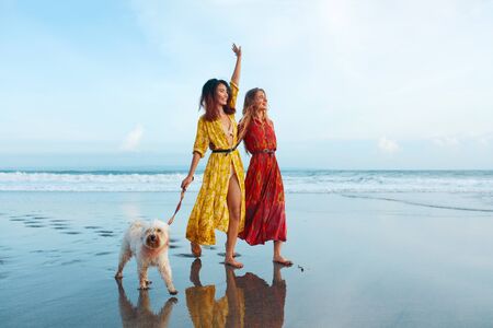 Beach. Women With Dog On Sandy Coast. Fashion Girls In Bohemian Clothing Walking Barefoot With Pet On Tropical Shore. Boho Style For Fashion Look On Summer Vacation At Resort.の写真素材