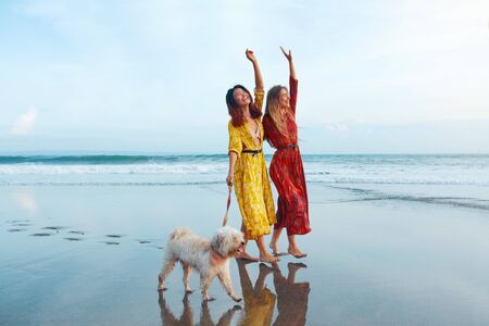 Women And Dog On Beach. Barefoot Girls With Pet On Dog-Friendly Coast. Happy Models Enjoying Walk With Cute Puppy On Summer Vacation At Tropical Ocean. Fashion Female In Trendy Bohemian Dresses.の写真素材