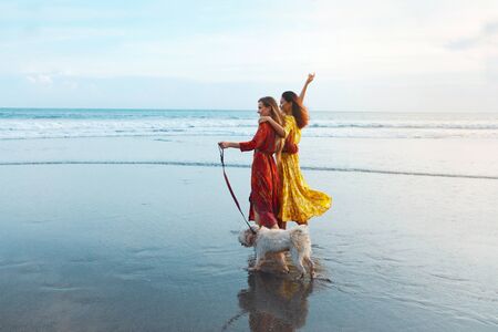 Women And Dog On Beach. Beautiful Girls Walking Barefoot With Pet On Dog-Friendly Coast. Fashion Models Enjoying Summer Vacation At Tropical Ocean. Fashion Female In Trendy Bohemian Dresses.の写真素材