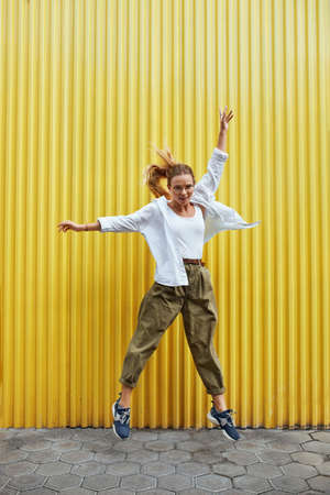 Urban. Fashion Girl Motion Portrait. Sexy Model In Casual Clothes And Glasses Against Yellow Iron Fence. Beautiful Woman In White Shirt And Top Tank Jumping On Pavement Near High Wall.の写真素材