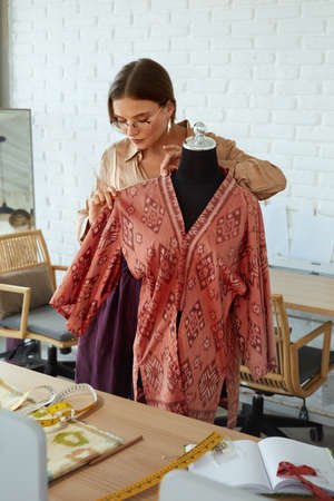 Fashion Designer At Workshop. Happy Woman In Stylish Clothes Working At Atelier. Success Self Employed Tailor Keeping Hand On Mannequin And Improving Design Of Elegant Dress.の写真素材