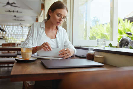 Business. Woman At Cafe Working Remotely. Girl In Casual Clothes And Glasses With Laptop Writing Something In Notebook. Digital Technologies For Creative Job At Comfortable Place On Summer Vacation.の写真素材