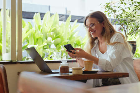 Online Job. Girl Works On Laptop And Using Smartphone At Cafe. Beautiful Business Woman In Casual Clothes And Glasses Portrait. Digital Technologies For Working, Shopping And Studying.の写真素材