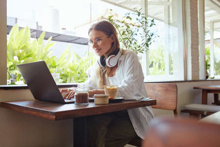 Woman. Online Work At Coffee Shop. Happy Girl In Casual Clothes With Laptop And Headphones Typing Something. Modern Digital Technologies For Remote Job Or Education At Comfortable Workplace.の写真素材