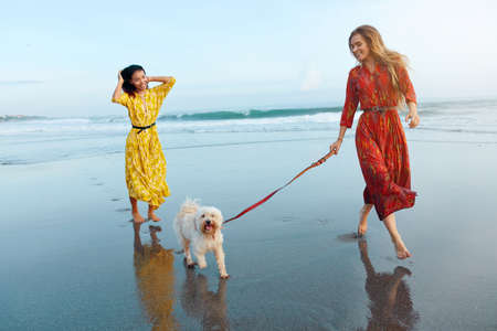 Girls With Dog On Beach. Barefoot Women In Maxi Boho Dresses Enjoying Summer Vacation With Pet Near Tropical Ocean. Beautiful Models In Trendy Outfit On Dog-Friendly Coast Near Sea.の写真素材
