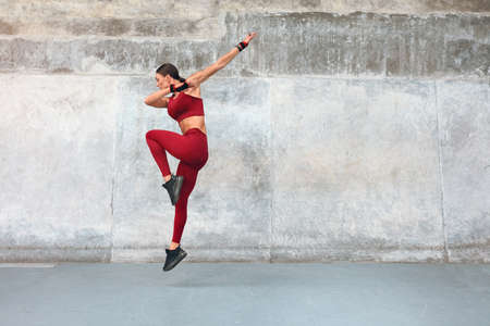Fitness. Woman Doing Jumping Workout At Outdoor Stadium. Full-Length Portrait Of Sporty Girl With Strong Muscular Body In Fashion Sportswear Doing Intense Cardio Exercise Against Concrete Wall.の写真素材