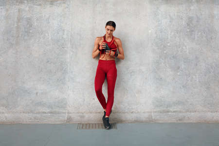 Gym. Fitness Girl With Bottle Of Protein Shake And Resistance Band Standing Near Wall. Sporty Woman In Fashion Sportswear Between Intense Cardio Workout. Outdoor Exercising As Urban Lifestyle.の写真素材
