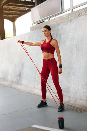 Sport. Woman With Resistance Band Exercising Outdoor. Fitness Girl In Fashion Sportswear Working Out Against Urban Background. Protein Shake And Cardio Training For Strong Muscular Body.の写真素材