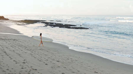 Surfing. Girl With Surfboard On Sandy Beach. Female Surfer Carrying Board And Going To Surf From Surf Spot In Bali. Water Sport On Summer Vacation For Active Lifestyle.の写真素材