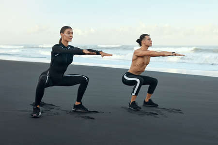 Sport. Man And Woman Squatting On Beach. Couple Of Young Sporty People In Fashion Fitness Outfit Warming Up Before Workout. Outdoor Morning Training For Strong Muscular Body.の写真素材