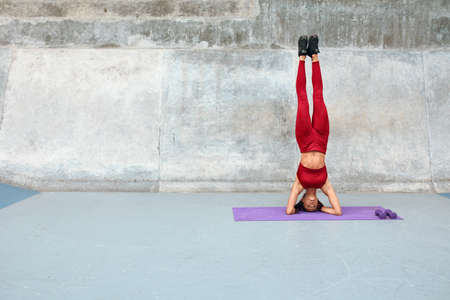Workout. Fitness Girl Standing On Head. Sexy Woman With Raising Legs Exercises On Yoga Mat At Outdoor Stadium. Fit Female With Sexy Muscular Body In Fashion Sportswear Training Against Concrete Wall.の写真素材