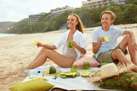 Summer. Family Picnic On Beach. Portrait Of Young Woman With Son Enjoying Lunch At Tropical Ocean. Mother And Handsome Boy On Vacation Near Sea. Organic Fruit And Natural Vitamins.の写真素材
