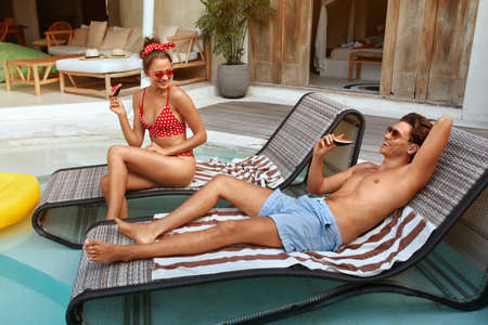 Summer Vacation At Pool. Couple Eating Watermelon And Enjoying Summertime At Tropical Villa. Handsome Man And Woman In Fashion Swimsuit Relaxing On Poolside. Honeymoon Or Romantic Weekend At Resort.の写真素材