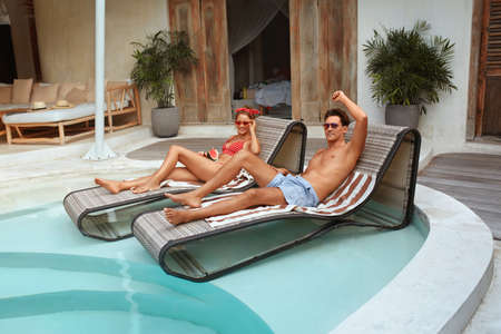 Man And Woman At Pool. Couple In Love Resting On Poolside At Tropical Villa. Handsome Guy And Sexy Girl In Fashion Swimsuit Enjoying Summer Vacation, Honeymoon Or Romantic Weekend At Resort.の写真素材