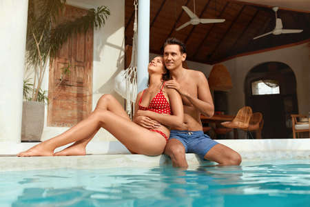 Man And Woman At Pool. Couple In Love Sitting On Poolside And Enjoying Summer Vacation At Tropical Villa. Handsome Guy Hugs Beautiful Girl In Fashion Swimsuit. Romantic Weekend At Resort.の写真素材