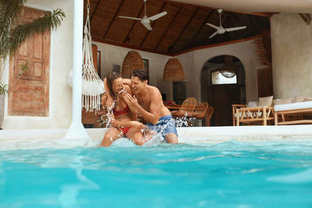 Couple In Love At Pool. Handsome Man And Beautiful Woman Sitting On Poolside, Splashing Water And Enjoying Summer Vacation. Fashion Male And Female Having Fun On Honeymoon At Tropical Villa.の写真素材