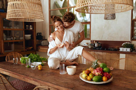 Healthy Diet. Couple Cooking Fresh Organic Food For Lunch At Kitchen In Tropical Villa. Man Hugs Woman Sitting On Wooden Table. Natural Vitamins For Nutrition On Summer Vacation Or Romantic Weekend.の写真素材