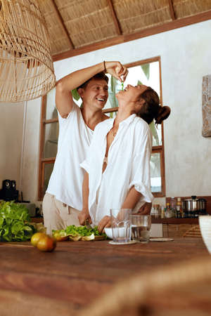 Couple At Kitchen. Handsome Man Feeding Beautiful Woman While She Cutting Fresh Organic Vegetables For Salad. Healthy Diet On Romantic Weekend, Honeymoon Or Summer Vacation At Tropical Villa.の写真素材
