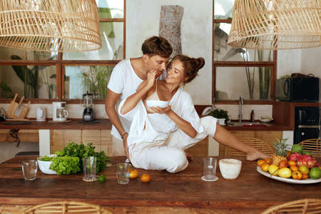 Lunch. Couple Cooking At Kitchen On Romantic Weekend. Beautiful Woman Sitting On Wooden Table Feeding Happy Man. Fresh Organic Vegetables And Natural Vitamins For Healthy Lifestyle.の写真素材