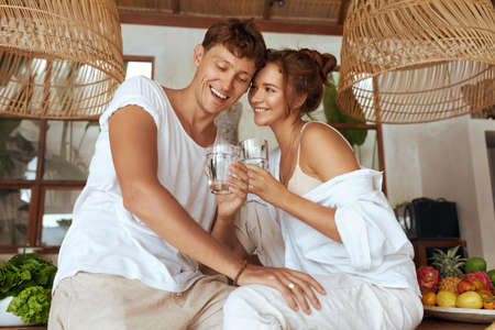 Couple At Kitchen Drinking Water. Man And Woman Holding Glasses, Hugging And Having Fun. Healthy Diet And Hydrate Recovery On Weekend, Honeymoon Or Summer Vacation At Tropical Villa.の写真素材