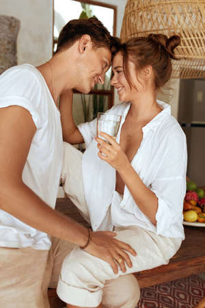 Romantic Couple Drinking Water At Kitchen. Beautiful Woman And Handsome Man Sitting On Wooden Table And Looking Lovely. Healthy Diet And Hydrate On Summer Vacation At Tropical Villa.の写真素材