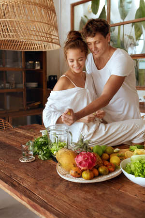 Healthy Diet. Romantic Couple Cooking Vegetarian Food At Kitchen In Tropical Villa. Woman Sitting On Table And Man Squeezing Lemon For Detox Cocktail Or Smoothie. Natural Vitamins For Nutrition.の写真素材