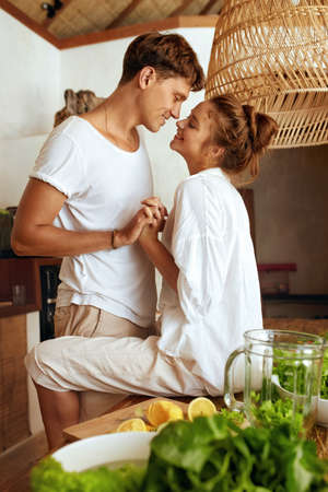 Lifestyle. Romantic Couple At Kitchen Cooking Healthy Vegetarian Food. Woman Sitting On Wooden Table And Standing Man Bonding Hands. Enjoying Romantic Weekend Or Honeymoon At Tropical Villa.の写真素材