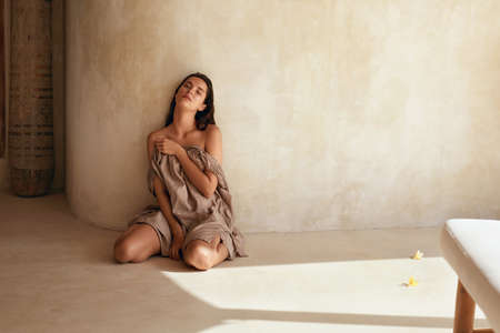 Woman. Portrait Of Beautiful Model Sitting Near Wall On Terrace. Seductive Brunette With Perfect Smooth Tanned Skin Posing On Floor With Flowers In Sunlight And Looking At Camera.の写真素材