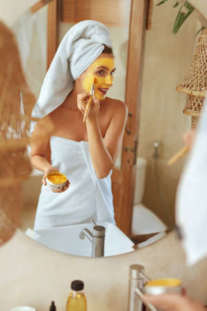 Woman Applying Face Mask. Wrapped In Bath Towel Model Standing In Bathroom And Looking At Mirror. Facial Skin Covered With Beauty Product. Daily Routine For Healthy Derma.の写真素材