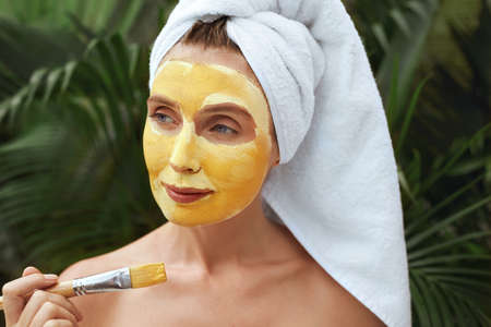 Woman Applying Clay Mask With Brush. Model In Bath Towel Using Skin Care Product For Oil Derma. Portrait Of Female Against Tropical Greenery Background At SPA Resort.の写真素材