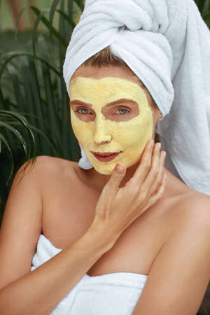 Woman In Bath Towel With Turmeric Mask On Face. Close Up Portrait Of Young Female Touching Facial Skin Covered With Beauty Product. Model After SPA Treatment Against Tropical Greenery.の写真素材