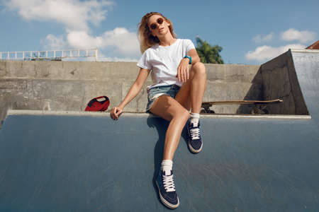 Summer. Skater Girl Sitting On Concrete Skate Ramp At Skatepark. Female Teenager In Casual Outfit With Skateboard. Urban Sport As Lifestyle For Active Teens In City.の写真素材