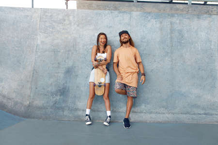 Skaters. Girl And Guy At Skatepark. Portrait Of Hipster Couple In Casual Outfit With Skateboard Against Concrete Wall At Park. Urban Subculture Style And Summer Sport In City. Active Lifestyle.の写真素材
