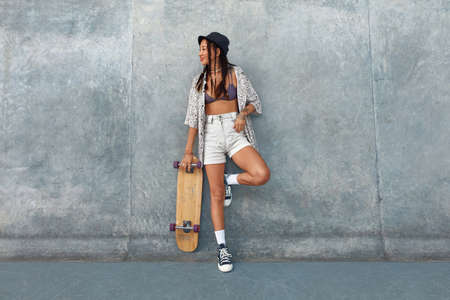 Skater Girl At Skatepark. Full-Length Portrait Of Female Hipster In Casual Outfit With Skateboard Against Concrete Wall At Skate Park. Urban Subculture Style And Summer Sport In City. Active Lifestyle.の写真素材