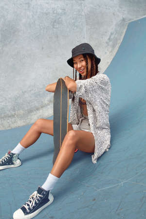 Skateboarding. Asian Girl In Urban Outfit With Skateboard Sitting On Ramp Against Concrete Wall At Skatepark. City Sport As Hobby For Active Teens. Skater Subculture And Summer Activity As Lifestyle.の写真素材