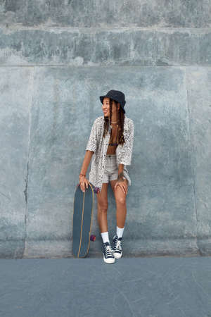 Skater Girl At Skatepark. Full-Length Portrait Of Female Hipster In Casual Outfit With Skateboard. Teenager Looking Away Against Concrete Wall At Skate Park. Urban Subculture Style And Summer Sport In City.の写真素材