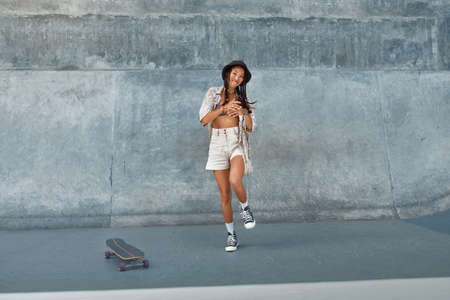 Portrait Of Skater Girl In Skatepark Having Fun. Female Teenager In Urban Outfit With Skateboard Against Concrete Wall. Summer Skateboarding With Modern Sport Equipment As Part Of Active Lifestyle.の写真素材