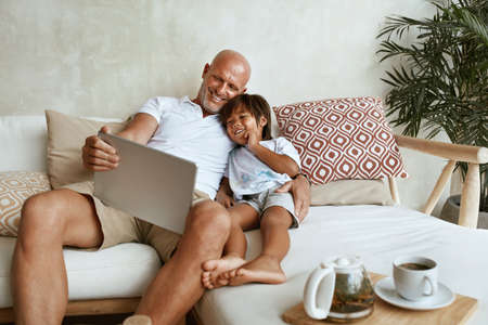 Father And Son With Tablet Portrait. Mixed Race Family Of Dad And Little Boy Using Portable Digital Device Sitting On Sofa Together. Technology For Education And Fun At Tropical Resort.の写真素材