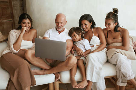 Big Family With Laptop On Sofa. Mixed Race People Using Portable Computer, Drinking Tea And Enjoying Weekend Together. Different Ethnic Parents With Daughters And Little Boy At Home.の写真素材