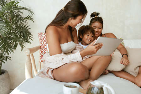 Siblings Using Tablet At Home. Portrait Of Different Age And Gender Asian Children Sitting On Sofa With Portable Digital Device. Education On Family Summer Vacation With Sisters And Little Brother.の写真素材